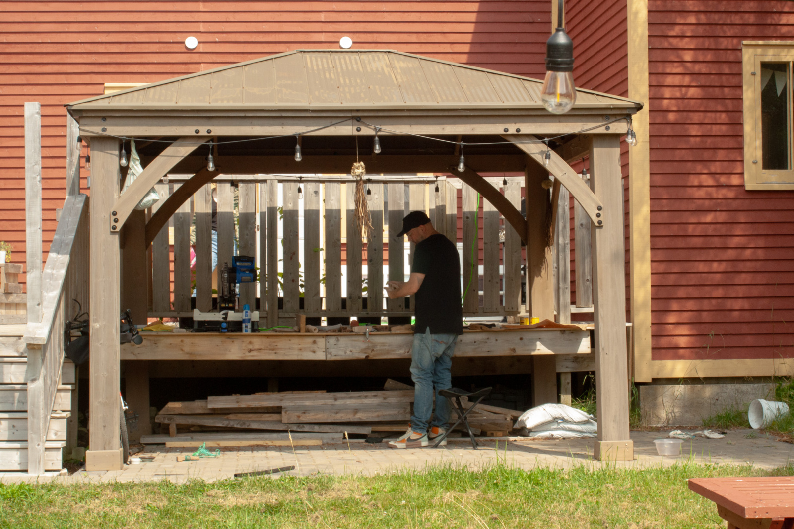 A artist working in the outdoor maker's space, covered by an awning, casting shade on the worktable. The patio leading to the gallery is visible behind the awning, and grass is visible outwards from the maker's space.